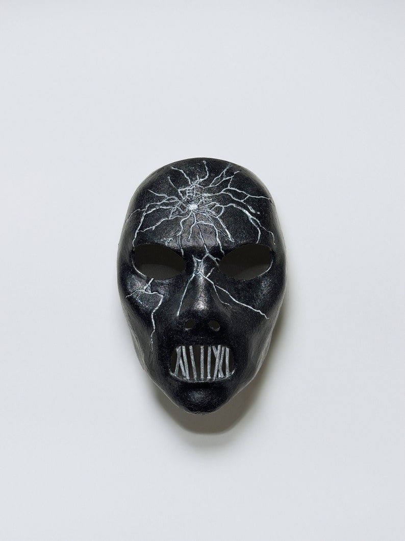 May include: A black mask with a cracked design and white lines. The mask has a mouth with white lines that resemble teeth.