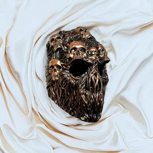 May include: A bronze-colored skull mask with multiple skulls sculpted on the surface. The mask has large eye sockets and a textured, organic design. The skulls are detailed with gold accents, creating a striking contrast.