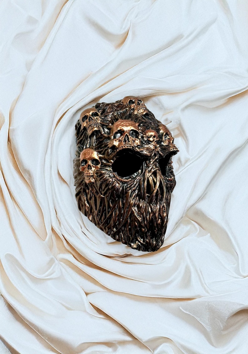 May include: A bronze-colored skull mask with multiple skulls sculpted on the surface. The mask has large eye sockets and a textured, organic design. The skulls are detailed with gold accents, creating a striking contrast.