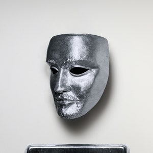 May include: A silver mask with a human face, the eyes are hollow and the mouth is slightly open. The mask is on a white background.
