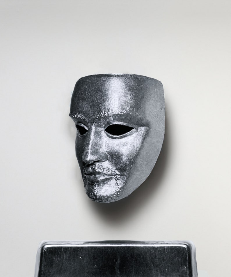 May include: A silver mask with a human face, the eyes are hollow and the mouth is slightly open. The mask is on a white background.