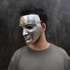 May include: A person wearing a silver mask that covers the top half of their face. The mask has a realistic human face sculpted on it. The person is wearing a gray t-shirt.