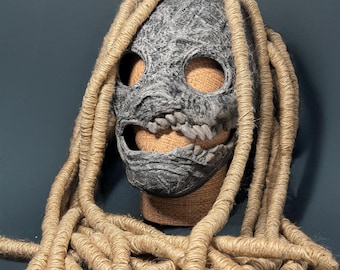 May include: A gray and black mask with a toothy grin, featuring a textured surface and a design reminiscent of a skull. The mask is adorned with long, thick, brown dreadlocks.