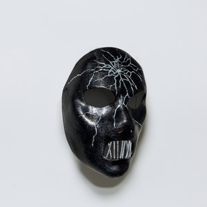 May include: A black mask with a white cracked design and a white metal mouth guard. The mask is made of a smooth, hard material.
