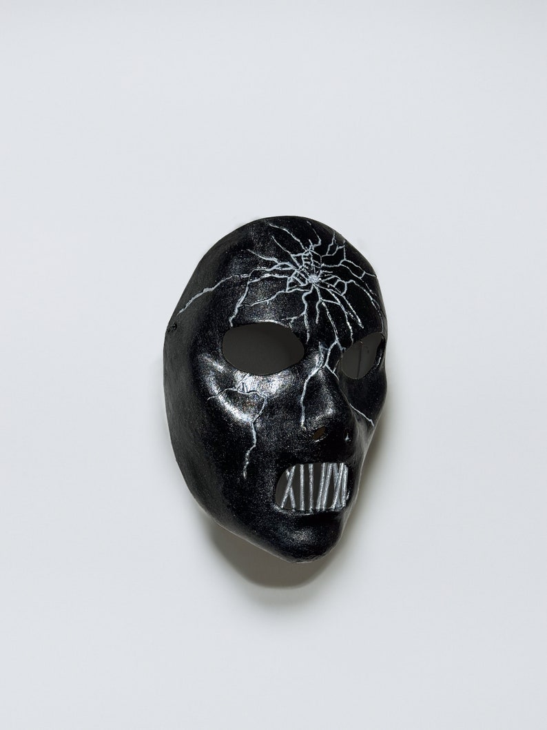 May include: A black mask with a white cracked design and a white metal mouth guard. The mask is made of a smooth, hard material.