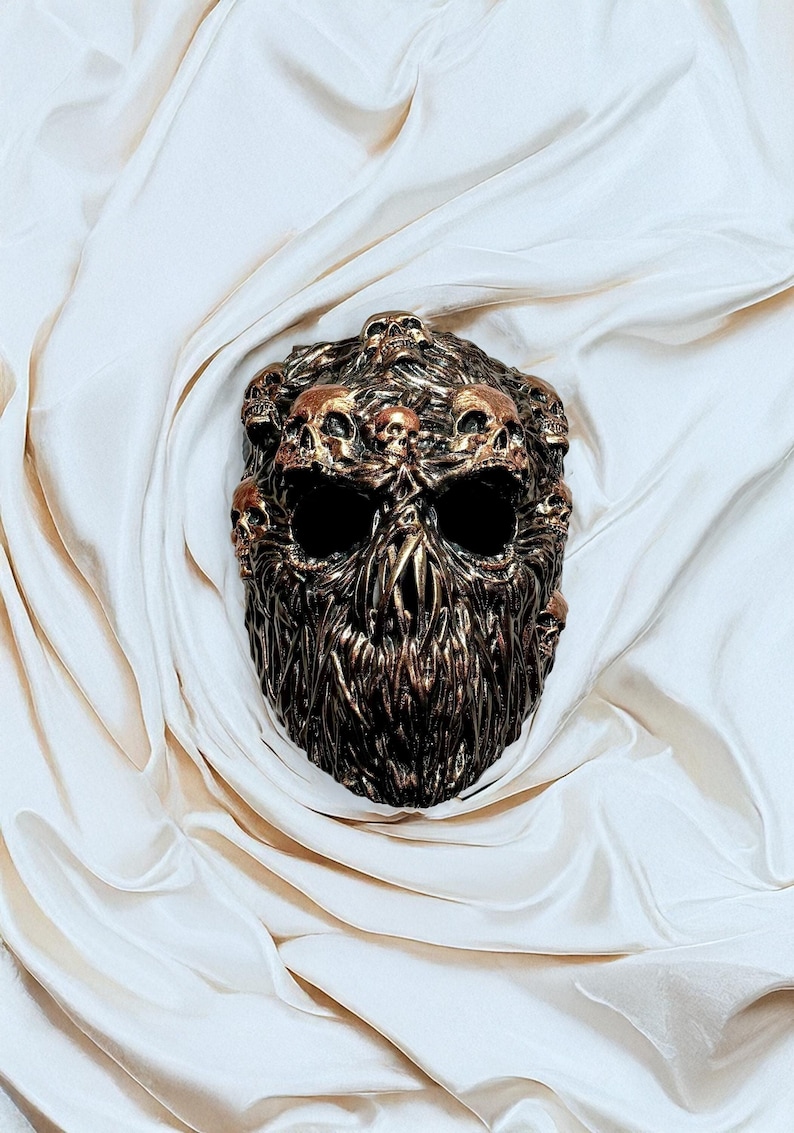 May include: A bronze-colored skull mask with multiple smaller skulls on top. The mask has large eye sockets and a textured, beard-like lower section. The mask is displayed on a white, draped fabric.