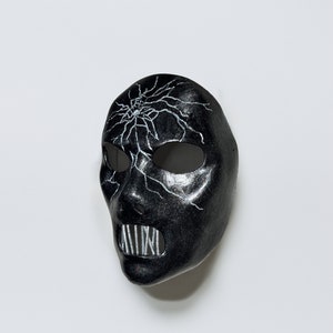 May include: A black masquerade mask with a white crackle design and a white, toothy mouth.