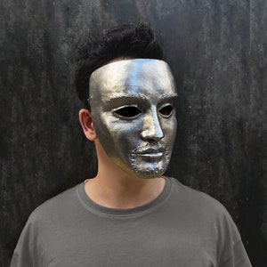May include: A silver face mask with a detailed design. The mask covers the forehead, eyes, nose, and mouth. The mask is made of a smooth, metallic material.