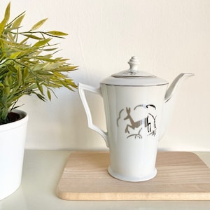 May include: White ceramic teapot with a silver deer design. The teapot has a silver lid and a spout. The teapot is sitting on a wooden surface.
