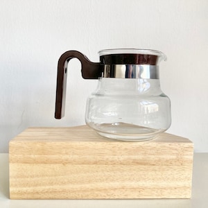 Può includere: Una caffettiera in vetro trasparente con un manico in plastica marrone e una fascia metallica argentata intorno alla parte superiore. La caffettiera è appoggiata su una superficie in legno.