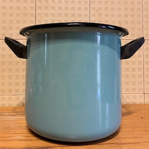 Op de afbeelding: Een lichtblauwe emaille pot met zwarte randen en handvatten. De pot heeft een cilindrische vorm en staat op een houten oppervlak. Het is een keukenartikel.