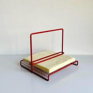 May include: A red wire napkin holder with a rectangular base and a vertical handle. The holder contains a stack of light yellow napkins. The design is simple and modern, suitable for a kitchen or dining area.