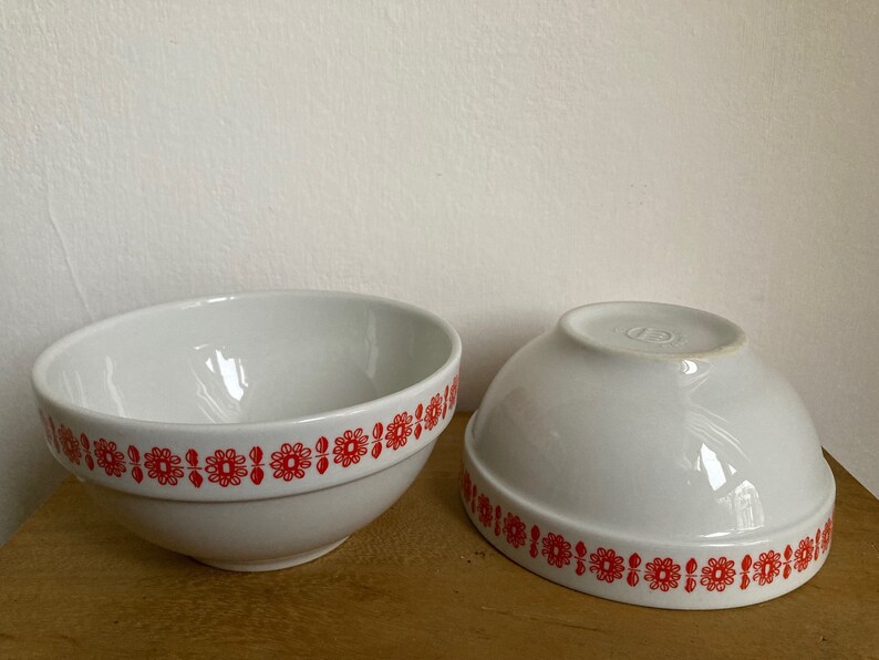May include: Two white ceramic bowls with a red floral pattern around the rim. The bowls are empty and sitting on a wooden surface.