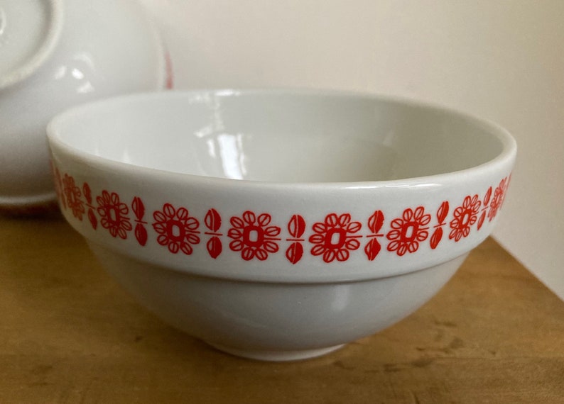 May include: White ceramic bowl with a red floral pattern around the rim. The bowl is empty and sitting on a wooden surface.