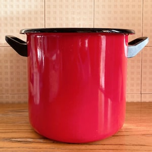 Può includere: Una pentola in smalto rosso vivo con bordo e manici neri. La pentola ha una finitura lucida ed è progettata per cucinare. I manici sono neri e argentati. La pentola è su una superficie di legno.