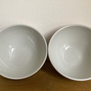 May include: Two white ceramic bowls, one on the left and one on the right, are shown on a wooden surface. The bowls are round and have a smooth, glossy finish.