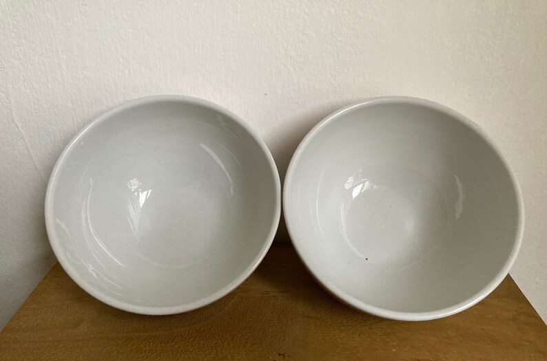 May include: Two white ceramic bowls, one on the left and one on the right, are shown on a wooden surface. The bowls are round and have a smooth, glossy finish.