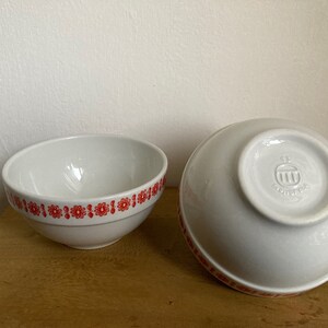 May include: Two white ceramic bowls with a red floral pattern around the rim. The bowl on the right has a small logo on the bottom.