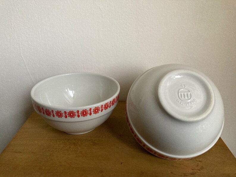 May include: Two white ceramic bowls with a red floral pattern around the rim. The bowl on the right has a small logo on the bottom.