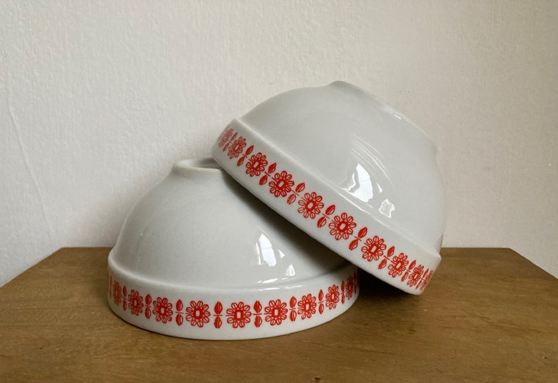 May include: Two white ceramic bowls with red floral designs around the rim. The bowls are stacked on top of each other.