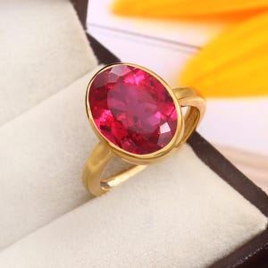 May include: A gold ring featuring a large, oval-shaped, red gemstone. The ring is displayed in a white velvet box.