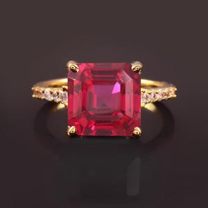 May include: A gold ring featuring a large, square-shaped, red ruby gemstone surrounded by smaller, clear gemstones.