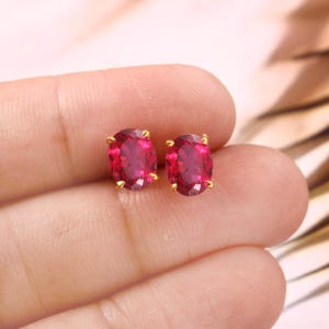 Puede incluir: Un par de pendientes de botón ovalados con piedras preciosas de color rojo rubí y garras doradas. Los pendientes se muestran sobre un fondo neutro, resaltando su color vibrante y diseño. Un artículo de joyería clásico.