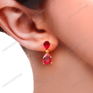 Puede incluir: Pendientes colgantes dorados con dos piedras preciosas rojas en forma de pera. Los pendientes tienen una pequeña piedra preciosa redonda en la parte superior y otra más grande en forma de lágrima en la parte inferior. Los pendientes se muestran junto a una oreja.