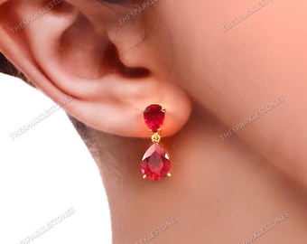 May include: Gold-toned drop earrings featuring two red pear-shaped gemstones. The earrings have a small, round gemstone at the top and a larger, teardrop-shaped gemstone below. The earrings are displayed next to an ear.