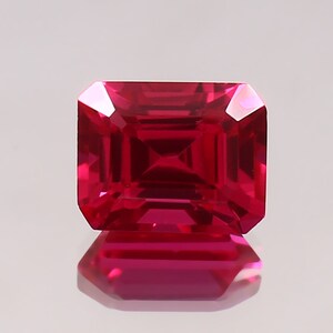 May include: A vibrant red emerald-cut gemstone with a high polish. The gemstone is reflecting light, creating a brilliant shine.