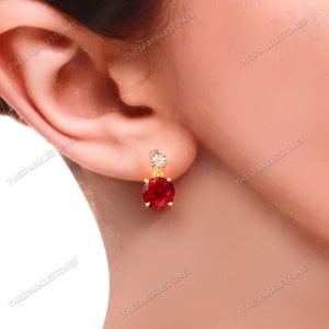 May include: A pair of gold earrings with a red gemstone and a small clear gemstone. The earrings are dangling and have a simple design.