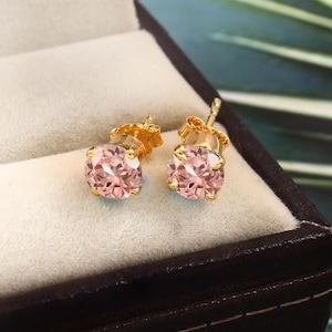 May include: A pair of gold stud earrings with pink round gemstones. The earrings are displayed in a small velvet box.