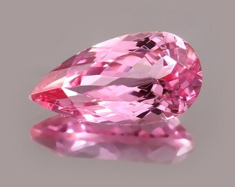 May include: A pear-shaped, faceted pink gemstone. The gem is a vibrant shade of pink, with a polished surface that reflects light. The gemstone is set against a neutral background, with its reflection visible below. The gem's facets catch the light.