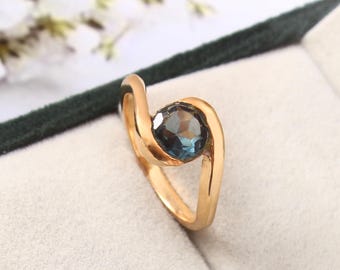 Könnte beinhalten: Ein goldfarbener Ring mit einem runden, facettierten, blauen Edelstein. Das Band des Rings umschließt den Edelstein in einem modernen, minimalistischen Design. Der Ring ist vor einem hellen Hintergrund platziert, um den Schmuck hervorzuheben.