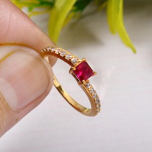 Può includere: Un anello d'oro con una singola pietra rossa quadrata e una fila di piccole pietre bianche attorno alla fascia.