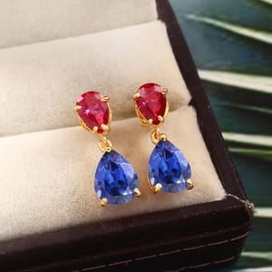 Puede incluir: Un par de pendientes de oro con piedras preciosas azules y rojas en forma de lágrima. Los pendientes tienen un diseño colgante con una piedra roja más pequeña en la parte superior y una piedra azul más grande en la parte inferior.