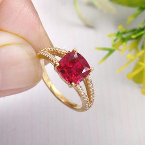 May include: A gold ring with a large, red, cushion-cut gemstone surrounded by smaller, clear gemstones.