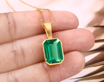 May include: A gold-colored necklace featuring a rectangular, emerald-green gemstone pendant. The pendant is set in a gold frame and hangs from a delicate gold chain. The gemstone has a faceted cut, reflecting light.