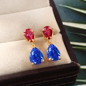 May include: A pair of gold-toned drop earrings featuring a red, teardrop-shaped gemstone at the top and a larger, blue, teardrop-shaped gemstone dangling below. The earrings are displayed in a jewelry box with a neutral-colored interior.