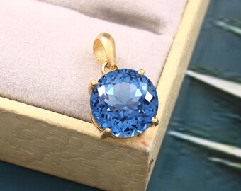 Puede incluir: Un colgante de oro con una gran piedra preciosa redonda azul. El colgante está engastado en un engaste de bisel de oro con un acabado liso y pulido.