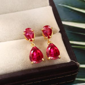 Peut inclure: Une paire de boucles d'oreilles en or avec deux pierres précieuses rouges en forme de goutte. Les boucles d'oreilles sont dans un écrin à bijoux.