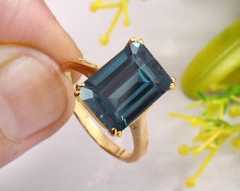 Könnte beinhalten: Ein goldener Ring mit einem großen, smaragdgeschliffenen blauen Edelstein. Der Ring ist auf einem schlichten Band gefasst.