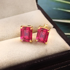 May include: A pair of gold earrings with square-shaped red gemstones. The earrings are in a small velvet box.