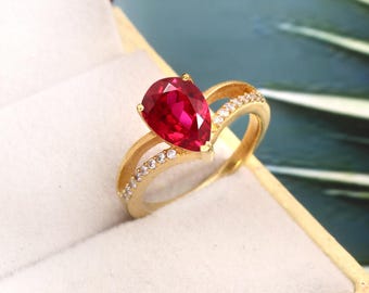 May include: A gold ring featuring a pear-shaped red gemstone and smaller clear gemstones set in a band.