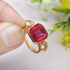 Puede incluir: Un anillo de oro con una piedra roja grande cuadrada y dos piedras transparentes más pequeñas en forma de pera.