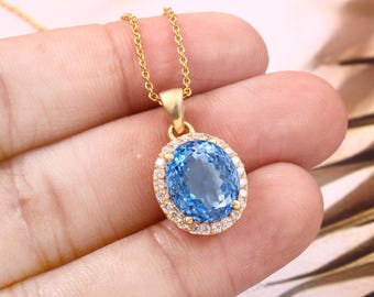 Puede incluir: Un collar de oro con un colgante que presenta una gran piedra de topacio azul ovalada rodeada por un halo de piedras preciosas más pequeñas y transparentes.