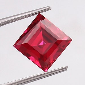 May include: A vibrant red square-shaped gemstone with a faceted cut. The gemstone is held by a pair of tweezers.