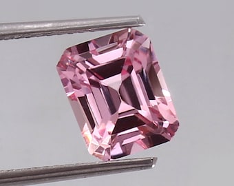 May include: A pink emerald cut gemstone with a faceted design. The gemstone is held in tweezers.