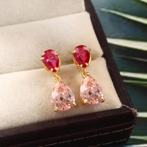 Puede incluir: Un par de pendientes de oro con piedras preciosas en forma de lágrima de color rosa y rojo. Los pendientes tienen un diseño colgante con una piedra roja más pequeña en la parte superior y una piedra rosa más grande en la parte inferior.