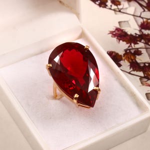 May include: A gold ring with a large, faceted, teardrop-shaped, deep red gemstone. The ring is set in a gold band and is presented in a white jewellery box. The gemstone's facets reflect light, creating a sparkling effect.